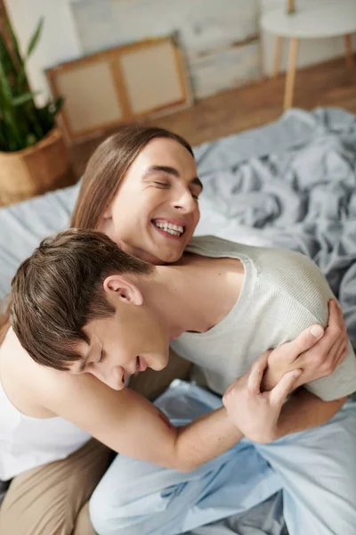 Vista ad alto angolo di uomo omosessuale allegro e dai capelli lunghi in pigiama da notte che abbraccia il fidanzato con gli occhi chiusi sul letto sfocato a casa al mattino — Foto stock