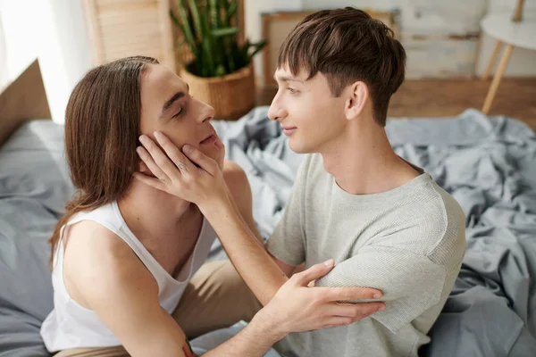 Vista lateral de jovem gay homem olhando e tocando rosto de longo cabelos namorado no pijama no borrado confortável cama em casa no manhã tempo — Fotografia de Stock