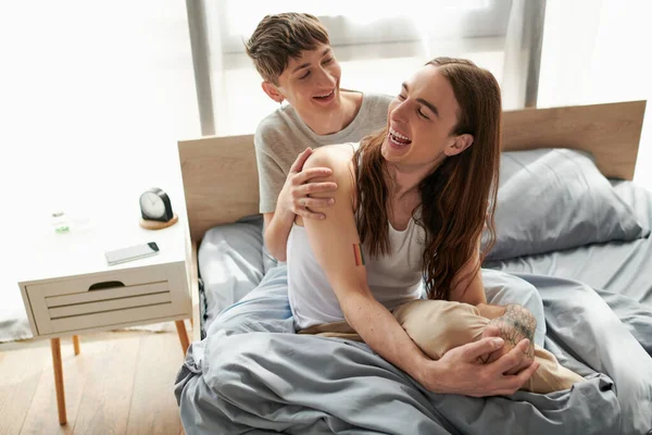 Alegre y joven pareja gay en pijama pasando tiempo juntos y hablando entre sí mientras descansan en una cómoda cama después de despertarse por la mañana en casa - foto de stock