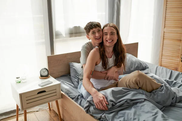 Junges lächelndes gleichgeschlechtliches Paar in Nachtwäsche, das sich umarmt und in die Kamera schaut, während es morgens auf dem Bett neben Smartphone und Wecker auf dem Nachttisch zu Hause liegt — Stockfoto