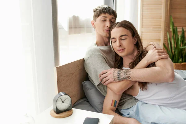Jovem gay homem com fechado olhos abraçando tatuado namorado no pijama enquanto sentado no cama perto de despertador e celular no cama mesa no manhã no casa — Fotografia de Stock