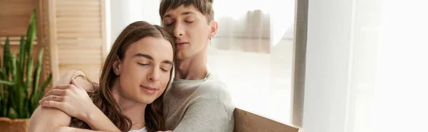 Jovem homossexual homem com olhos fechados abraçando o namorado de cabelos longos em roupas de dormir perto de cortinas com luz do dia em casa na hora da manhã, banner — Fotografia de Stock