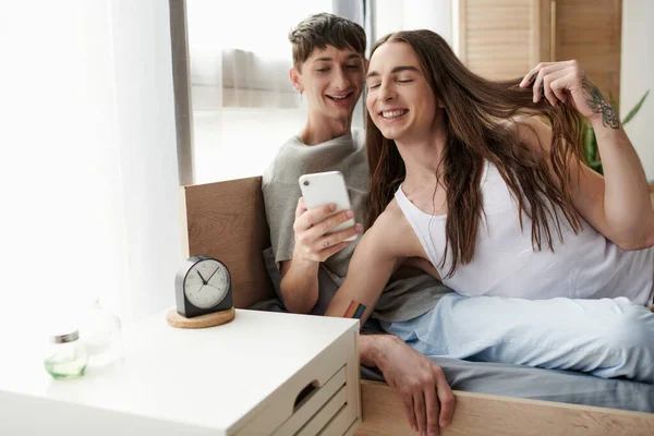 Smiling lgbt couple in sleepwear using mobile phone together while relaxing on cozy bed near alarm clock on bedside table in morning at home — Stock Photo