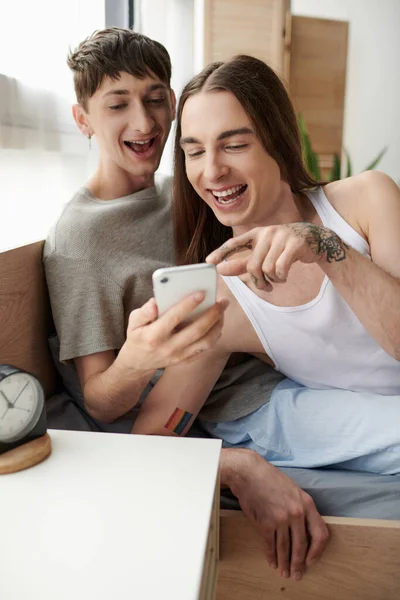 Alegre longo cabelos gay homem com tatuagem apontando para smartphone perto namorado no sleepwear sentado no cama ao lado de despertador no cama mesa no quarto no manhã — Fotografia de Stock