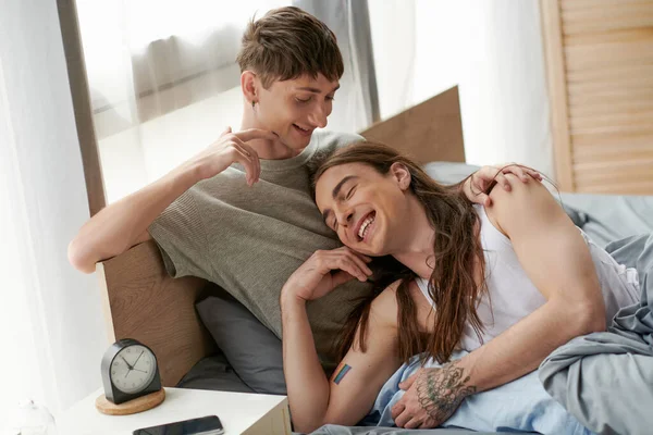 Positive homosexual man in pajama touching long haired and tattooed boyfriend on bed near cellphone and alarm clock on bedside table at home — Stock Photo