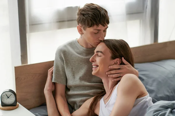 Молодой гей в сонной одежде целует счастливого длинноволосого парня с закрытыми глазами, сидя на кровати рядом с будильником на тумбочке дома утром — стоковое фото
