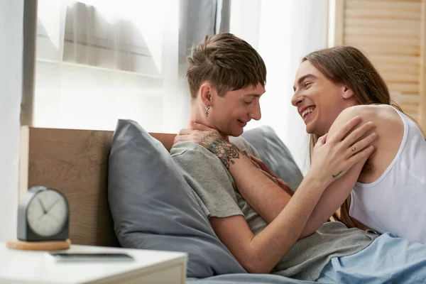 Positive same sex couple hugging and having conversation while relaxing on bed near blurred smartphone with blank screen on bedside table at home in morning time — Stock Photo