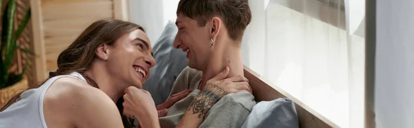 Щасливий і татуйований гей чоловік обіймає і дивиться на молодого хлопця в сплячому одязі, лежачи і сміючись разом на ліжку вдома вранці, банер — стокове фото