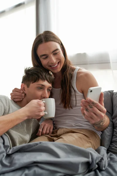 Hombre homosexual despreocupado y de pelo largo usando un teléfono inteligente y abrazando a su novio en pijama bebiendo café mientras están sentados juntos en una cómoda cama en casa por la mañana - foto de stock