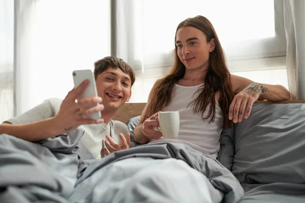 Беззаботный гей в пижаме держит мобильный телефон рядом с длинноволосым и татуированным парнем держа чашку кофе и сидя на кровати дома утром — стоковое фото
