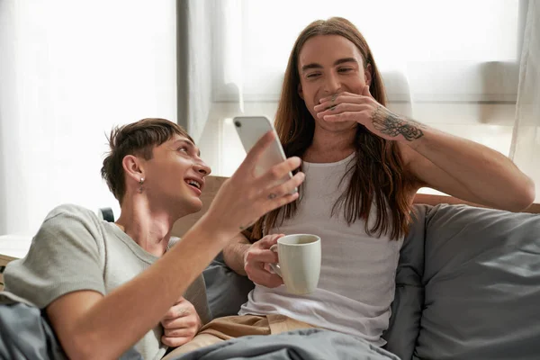 Alegre e jovem homossexual homem mostrando celular para feliz longo haired parceiro no pijama segurando xícara de café e descansando na cama no quarto moderno de manhã — Fotografia de Stock