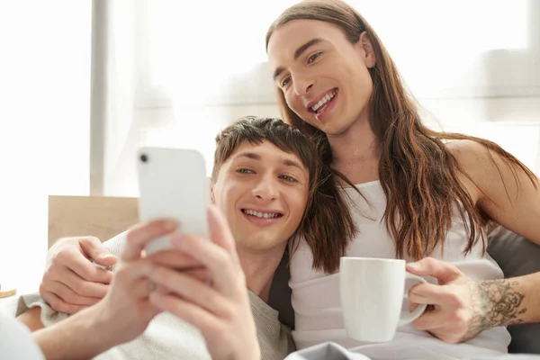 Jeune couple de même sexe en pyjama tenant une tasse de café et souriant tout en utilisant un smartphone flou et se reposant sur un lit confortable dans la chambre le matin à la maison — Photo de stock