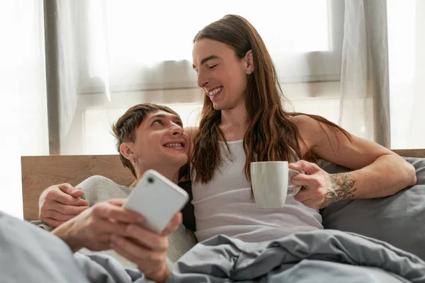 Positive homosexual man holding coffee cup while hugging and looking at boyfriend in pajama using blurred smartphone on comfortable bed at home in morning — Stock Photo