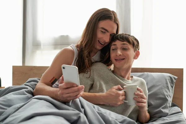 Sonriente y joven gay en pijama sosteniendo la taza de café y mirando el celular mientras se relaja en la cómoda cama en casa por la mañana - foto de stock