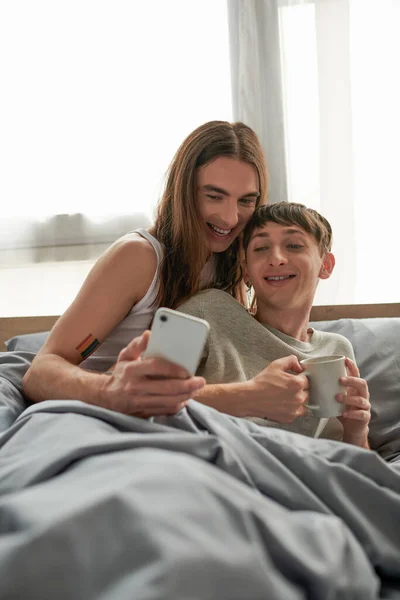 Joyeux homme gay aux cheveux longs et tatoué utilisant un smartphone à côté du jeune petit ami avec tasse de café tout en étant assis sous la couverture sur le lit à la maison le matin — Photo de stock