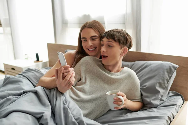 Улыбающийся длинноволосый гей, показывающий смартфон взволнованному парню в пижаме, держащему чашку кофе, лежа под одеялом на кровати дома утром — стоковое фото