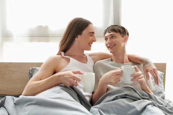 Smiling tattooed gay man in sleepwear hugging and looking at young happy boyfriend while holding coffee cup and resting on comfortable bed in morning at home — Stock Photo
