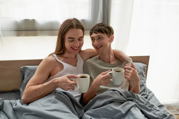 Улыбающийся длинноволосый и татуированный гей во сне держит чашку кофе и обнимает молодого бойфренда, просыпаясь утром дома на удобной кровати — стоковое фото
