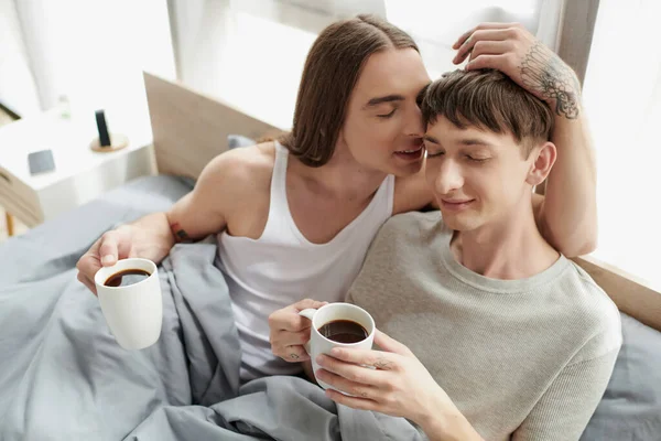 Cabello largo y hombre gay tatuado con los ojos cerrados hablando y tocando la cabeza del novio sonriente en ropa de dormir mientras sostiene la taza de café en la cama por la mañana en casa - foto de stock
