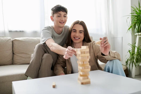 Jovem e feliz casal homossexual em roupas casuais sorrindo enquanto joga blocos de madeira jogo na mesa e passar o tempo juntos no apartamento moderno — Fotografia de Stock