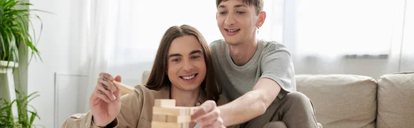 Junges und lächelndes gleichgeschlechtliches Paar in lässiger Kleidung, das unscharfe Holzklötze in der Nähe von Couch und verschwommene grüne Pflanzen im heimischen Wohnzimmer spielt, Banner — Stockfoto