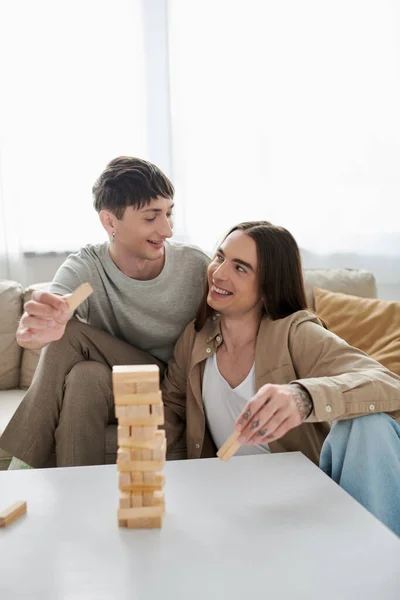 Young and cheerful same sex couple in casual clothes talking and looking at each other while holding parts of wood blocks game near table and couch at home — Stock Photo