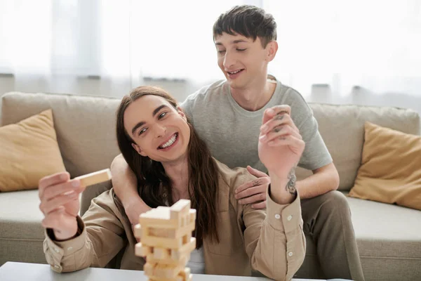 Smiling gay man hugging and looking at long haired and tattooed boyfriend while playing blurred wood blocks game on table near couch in living room at home — Stock Photo