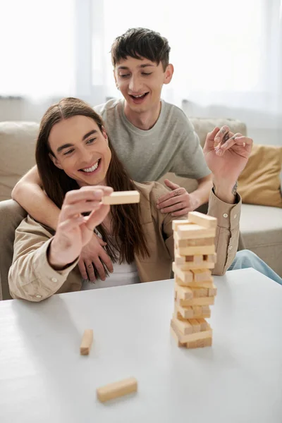 Positive gay man in casual clothes hugging tattooed and long haired boyfriend holding part of blurred wood blocks game on table in living room at home — Stock Photo