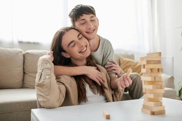 Young smiling gay man with closed eyes hugging long haired boyfriend in casual clothes near blurred wood blocks game on table in living room at home — Stock Photo