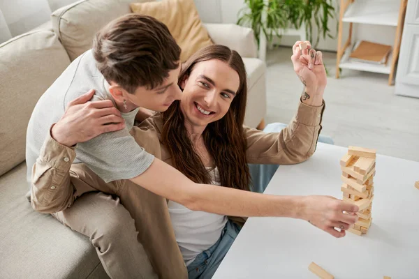 Positive long haired and tattooed homosexual man hugging boyfriend in casual clothes and playing wood blocks game on table while spending time at home — Stock Photo