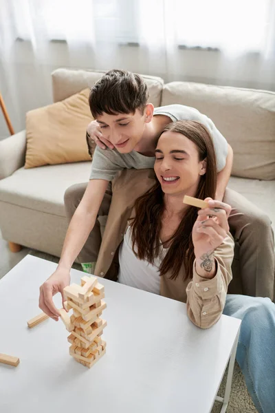 Jovem gay casal no casual roupas sorrindo enquanto jogar madeira blocos jogo no mesa perto confortável sofá enquanto passar tempo juntos no sala de estar no casa — Fotografia de Stock