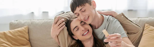 Young and positive same sex couple closing eyes while hugging and playing wood blocks game near couch in living room at home, banner — Stock Photo
