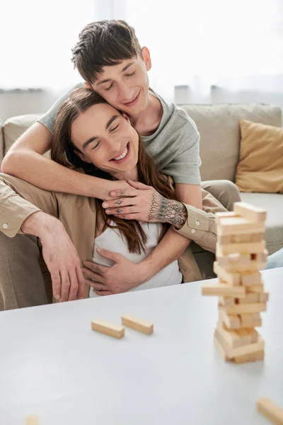 Smiling gay man with closed eyes hugging tattooed and long haired boyfriend in casual clothes near blurred wood blocks game and parts on table in living room at home — Stock Photo