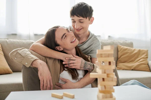 Smiling homosexual man hugging and looking at long haired boyfriend in casual clothes near blurred wood blocks game and parts on table in living room at home — Stock Photo