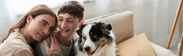 Hombre gay de pelo largo con tatuaje en la mano abrazando novio sonriente en ropa casual cerca del perro pastor australiano en un sofá moderno en la sala de estar en casa, pancarta - foto de stock