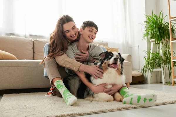 Young long haired gay man in socks and casual clothes hugging smiling boyfriend and petting Australian shepherd dog while spending time on carpet at home — Stock Photo