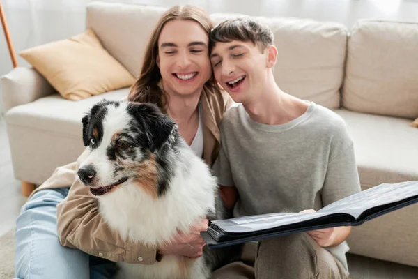 Alegre e jovem casal lgbt segurando álbum de fotos em mãos e olhando para cão pastor australiano bonito enquanto sentado ao lado do sofá na sala de estar moderna — Fotografia de Stock