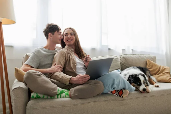 Cheerful lgbt couple in casual clothes looking at each other and using laptop while sitting together on comfortable couch with pillows near furry friend in modern living room — Stock Photo
