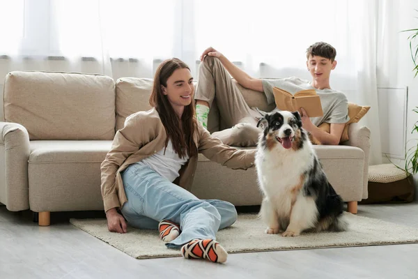 Счастливый гей читает книгу и лежит на удобном диване, в то время как его парень с длинными волосами играет с австралийской пастушьей собакой в свободное время дома — стоковое фото