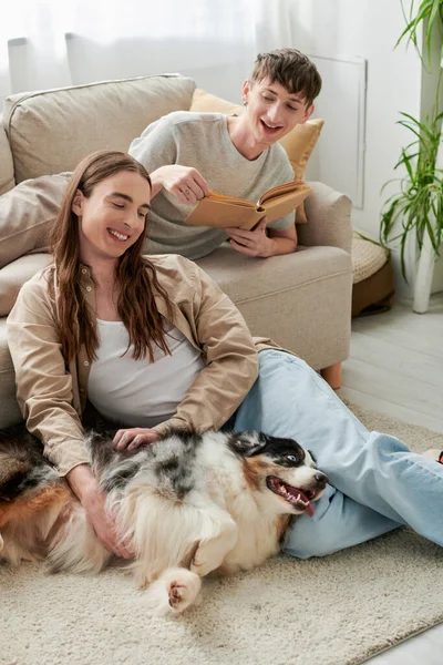 Alegre gay hombre con largo pelo abrazar australiano pastor perro mientras su tatuado novio en casual ropa descansando en sofá y celebración libro en moderno sala de estar - foto de stock