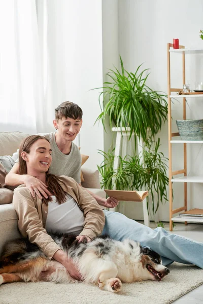 Fröhlicher schwuler Mann hält Buch in der Hand und ruht sich auf bequemer Couch aus, während er seinen Freund mit langen Haaren umarmt, der neben dem australischen Schäferhund in der modernen Wohnung auf dem Teppich sitzt — Stockfoto