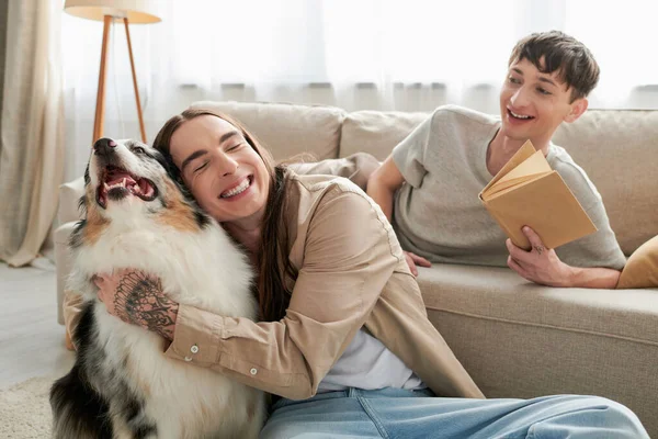 Happy gay man with tattoo smiling while hugging Australian shepherd dog next to cheerful gay man holding book and resting on comfortable couch in living room — Stock Photo