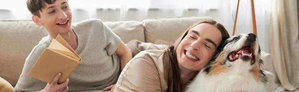 Happy gay man with tattoo smiling while hugging Australian shepherd dog next to cheerful gay man holding book and resting on comfortable couch in living room, banner — Stock Photo