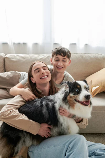 Alegre casal lgbt em roupas casuais sentado ao lado de sofá confortável e sorrindo juntos enquanto abraçando cão pastor australiano na sala de estar moderna — Fotografia de Stock