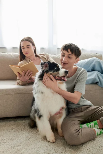 Young gay man with long hair reading book and resting on comfortable sofa next to his boyfriend with tattoo sitting on carpet with Australian shepherd dog in modern apartment — Stock Photo