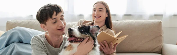 Happy young gay man with long hair holding book and resting on comfortable sofa next to his boyfriend cuddling Australian shepherd dog in modern apartment, banner — Stock Photo