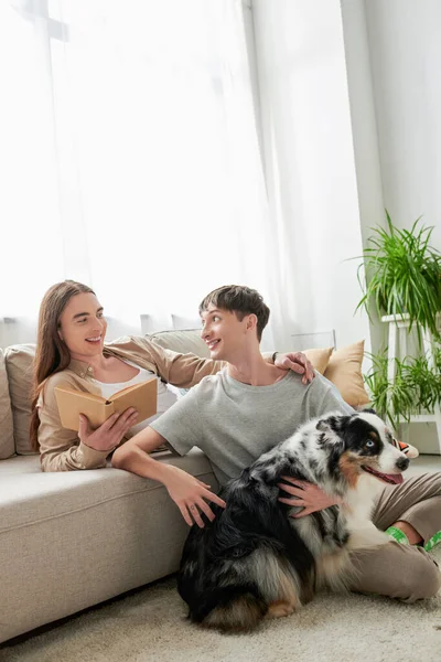 Happy gay man with long hair holding book and looking at tattooed and cheerful boyfriend hugging Australian shepherd dog in modern living room at home — Stock Photo
