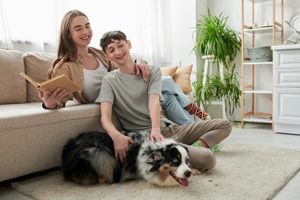 Cheerful gay man sitting on carpet and cuddling Australian shepherd dog young partner with long hair holding book and resting on sofa in modern living room — Stock Photo