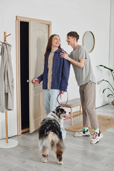 Pleased gay couple in casual outfits standing in modern hallway next to coat rack and holding leash with Australian shepherd dog and smiling together at home — Stock Photo