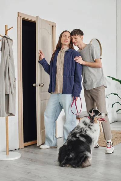 Young gay couple in casual outfits standing with closed eyes next to each other in hallway near coat rack and Australian shepherd dog with leash in modern apartment — Stock Photo